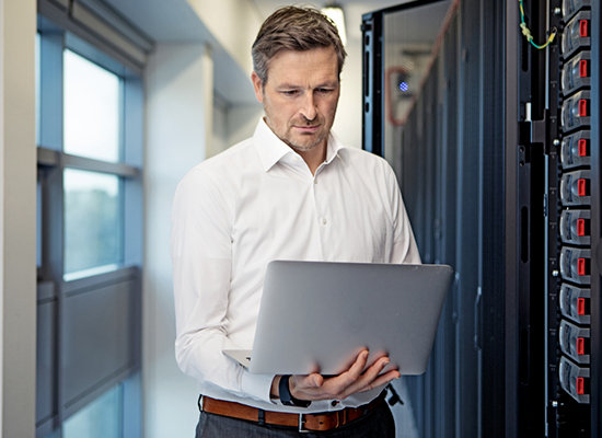 Man standing with white shirt holding laptop next to an open data center server